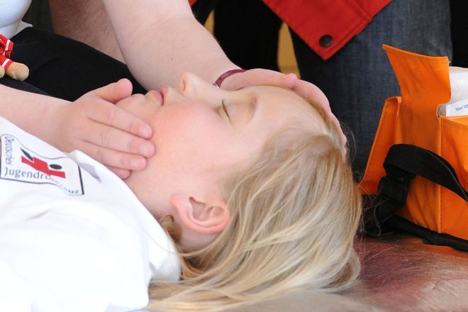 Foto: Bei einer Erste-Hilfe-Übung liegt ein Kind bewusstlos am Boden. Ein anderes Kind überprüft gerade die Atmung. Am Rande des Fotos ist eine Tasche mit Verbandsmaterial zu sehen.