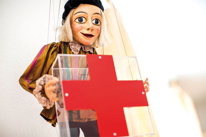 Foto: Eine junge DRK-Mitarbeiterin steht vor einer weißen Wand mit einem großen, roten Kreuz. Sie blickt direkt in die Kamera und wirkt insgesamt zuversichtlich und gelassen.