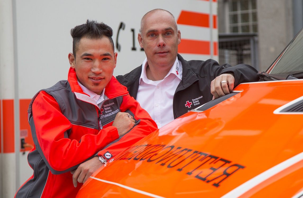 Foto: Eine junge Mitarbeiterin des DRK-Rettungsdienstes steht vor einem Rettungswagen und lacht in die Kamera. Sie wirkt insgesamt robust, warmherzig und aufgeschlossen.