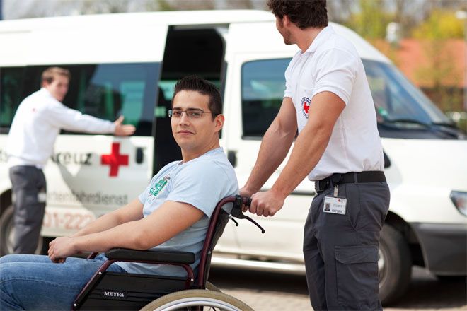 Foto: Ein junger Mann im Rollstuhl blickt zufrieden in die Kamera. Er wird von einem Mitarbeiter des DRK-Fahrdientes geschoben. Im Hintergrund öffnet ein weiterer Mitarbeiter die Schiebetür eines Transporters.