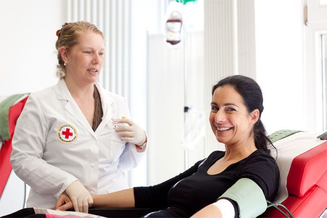 Foto: Eine Blutspenderin blickt entspannt und zuversichtlich in die Kamera. Währenddessen prüft eine DRK-Mitarbeiterin Blutdruck und Puls der Frau.