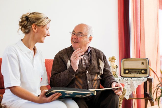 Foto: Eine DRK-Mitarbeiterin und ein älterer Herr sitzen auf einer Couch. Sie unterhalten sich, wobei das Strahlen in ihren Augen deutlich zu sehen ist.